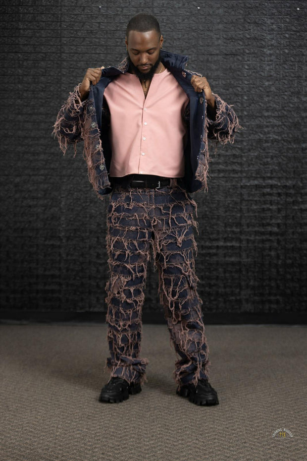 Man wearing a pink shirt, patterned jacket, and pants against a dark textured wall.